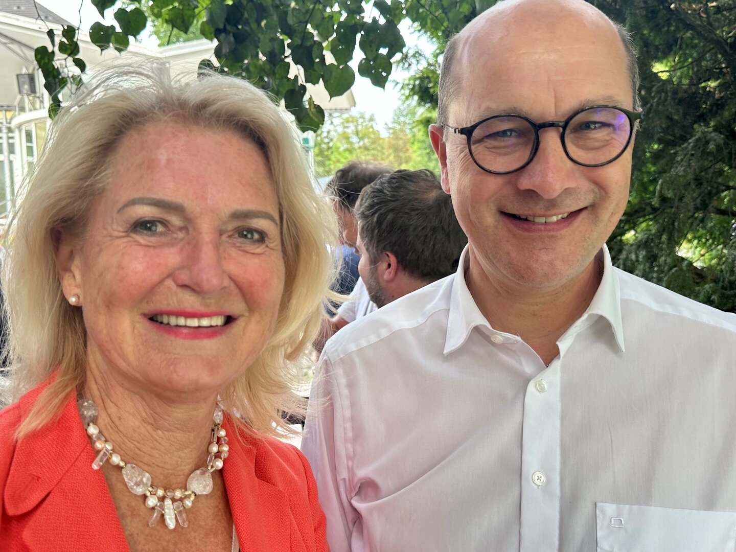 Doris Bures und Karl Nehammer lächeln in die Kamera, im Hintergrund Bäume und ein Gebäude.